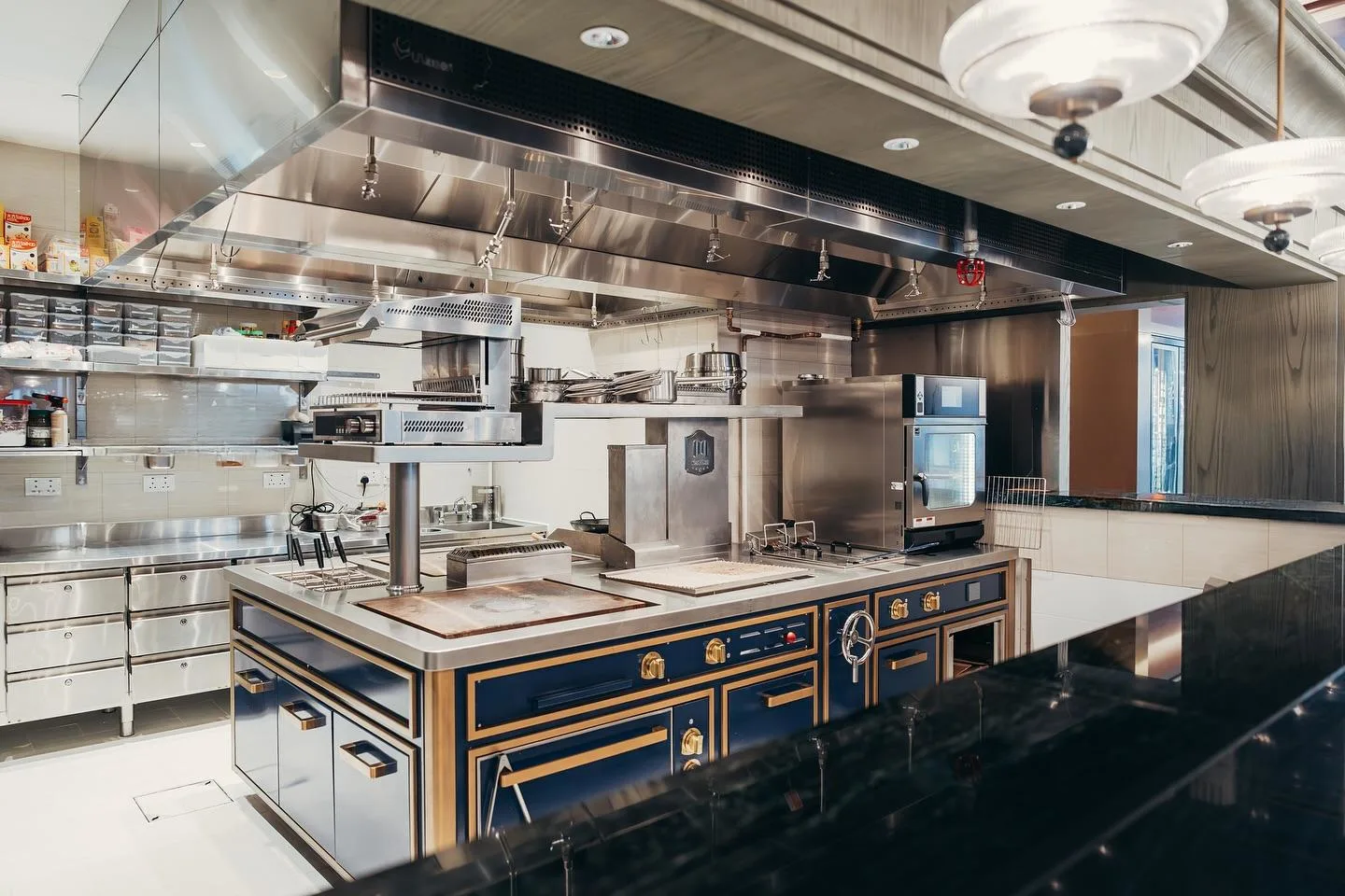 A professional hotel kitchen setup featuring blue cooking ranges and a large stainless steel overhead extraction hood.