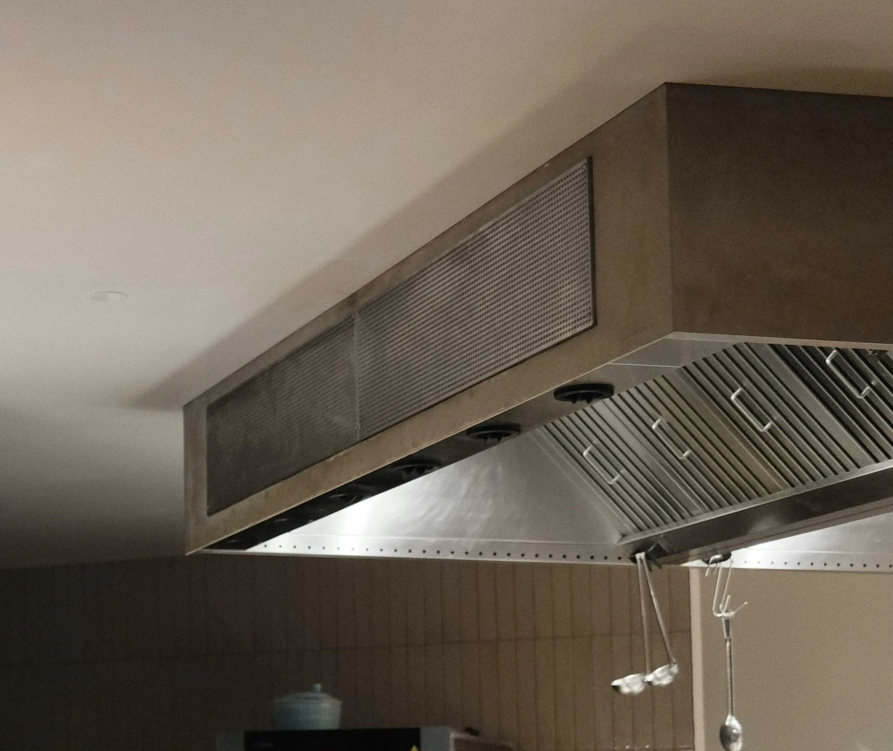 Close-up of a stainless steel commercial kitchen extraction hood featuring baffle grease filters and hanging utensils.