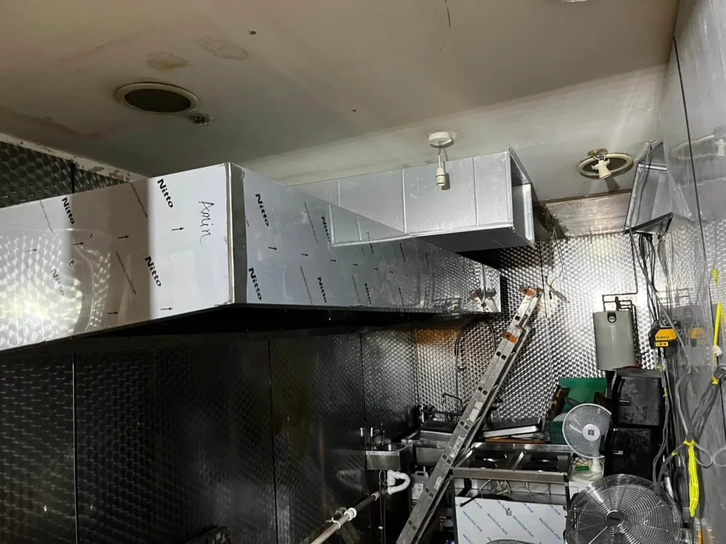 Stainless steel commercial kitchen extraction hood and ventilation ductwork being installed indoors.